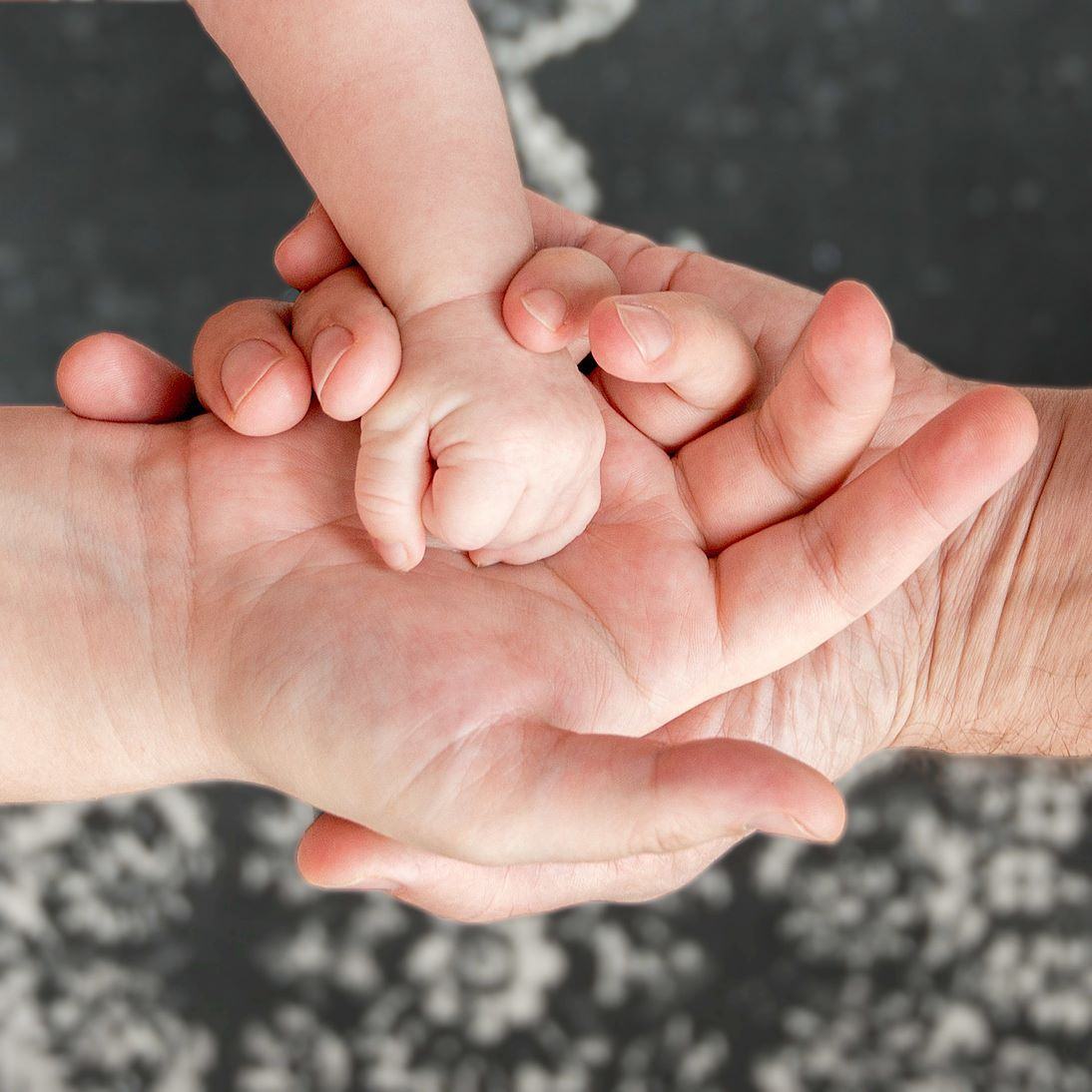 Mão de uma criança acolhida pelas mãos de dois adultos.