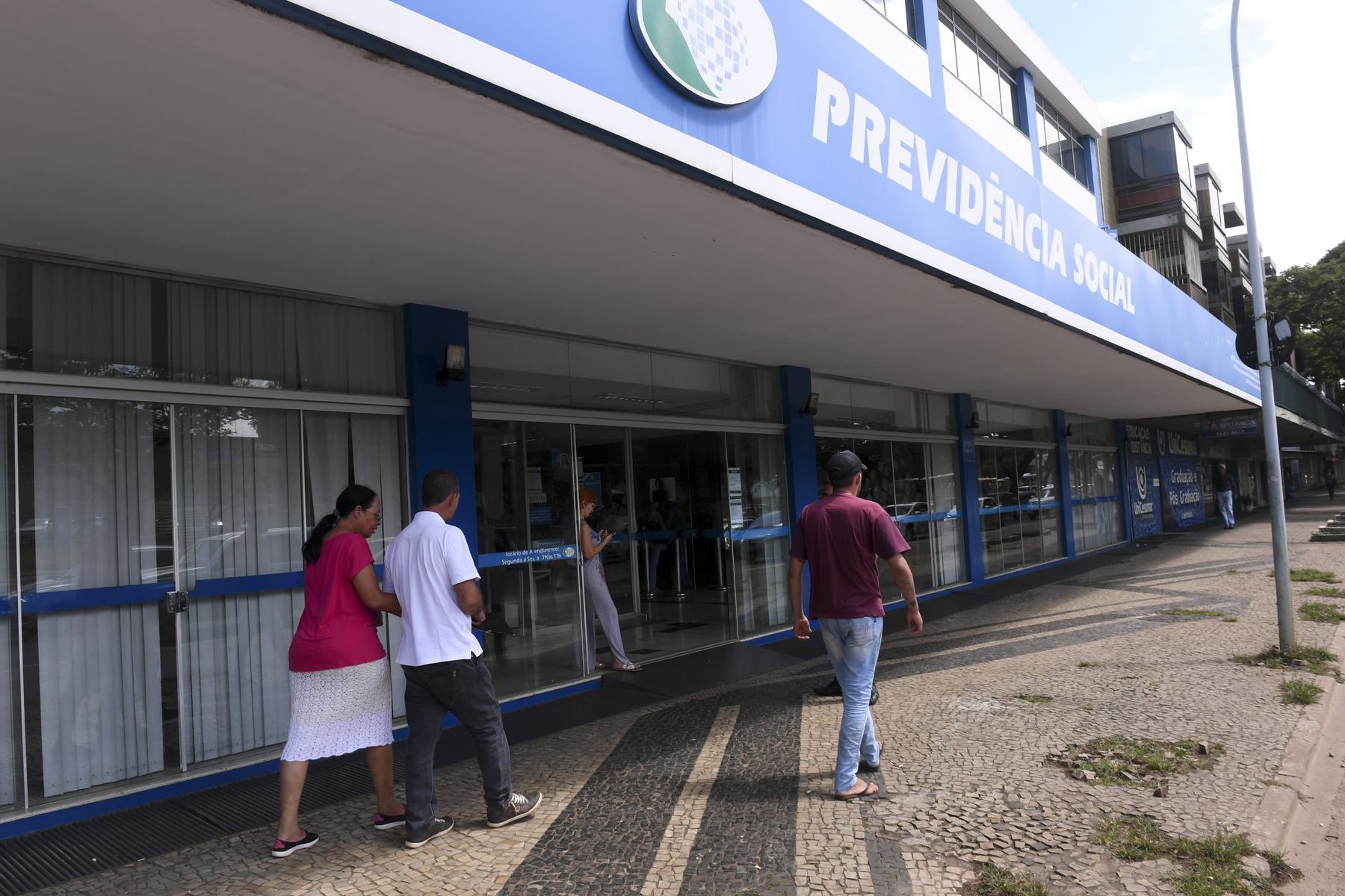 Fachada azul de agência do INSS com o letreiro Previdência Social e pessoas na entrada.